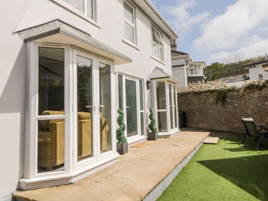 A patio area with stone tiles and artificial grass outside a two-story house with white framed glass doors and windows at Fonthill in Torquay