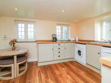 A kitchen with wooden floor a round table with flowers on it washing machine and coffee maker at The Linhay in Holsworthy