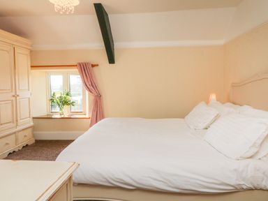 A bedroom with a white bed, a window with pink curtains, a flower vase on the window sill, and a wardrobe at The Linhay in Holsworthy