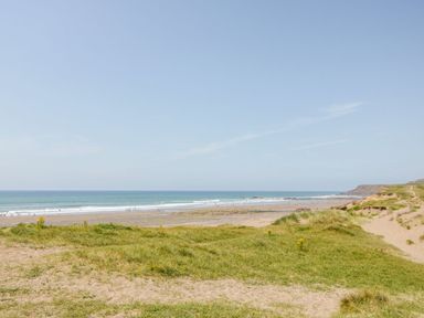 A sandy beach with grassy dunes and ocean waves under a clear sky at The Linhay in Holsworthy