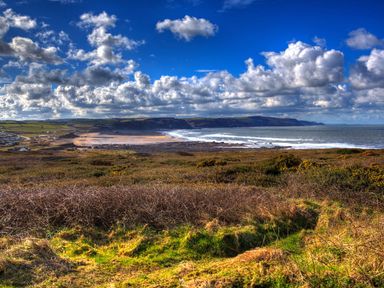 A coastal landscape with grassy and shrub covered land leading to a beach and ocean under a partly cloudy sky at The Linhay in Holsworthy