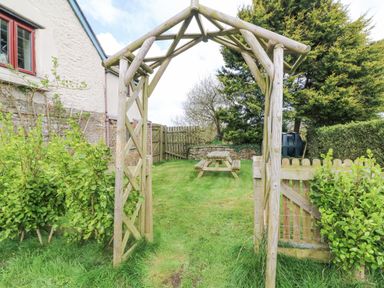 A wooden garden archway leading to a grassy yard with a wooden picnic table and a wooden fence at The Linhay in Holsworthy