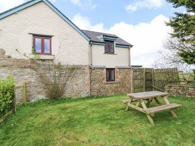 A grassy backyard with a wooden picnic table next to a stone and plaster house at The Linhay in Holsworthy