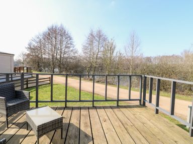 A wooden deck with a wicker chair and table overlooking a grassy area and trees at 109 Pentney Lakes in Pentney near King's Lynn