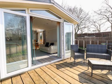 An outdoor wooden deck with wicker chairs and a table opening to a living room with a sofa and dining table at 109 Pentney Lakes in Pentney near King's Lynn