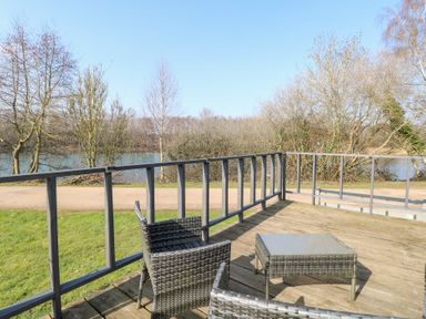 An outdoor deck with two wicker chairs and a glass-top table overlooking a lake and trees at 109 Pentney Lakes in Pentney near Kings Lynn