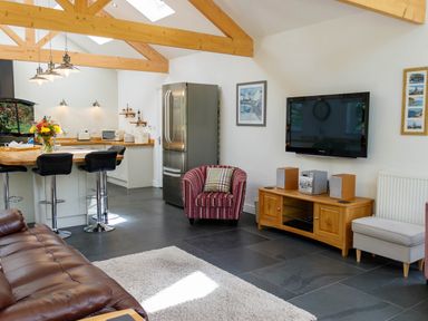 An open plan kitchen and living area with wooden beams black bar stools a red striped armchair and a wall mounted television at The Old Vicarage Retreat in Luxulyan
