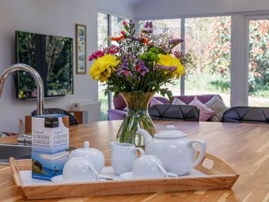 A wooden table with a tea set flowers and biscuits with chairs and a television in the background at The Old Vicarage Retreat in Luxulyan