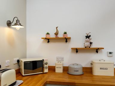 A kitchen countertop with a toaster microwave biscuit cake and bread containers and decorative shelves on white wall at The Old Vicarage Retreat in Luxulyan