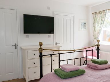 A bedroom with a metal bed frame folded towels on the bed a white dresser a wall-mounted TV a pink chair a window with patterned curtains and two white doors at The Old Vicarage Retreat in Luxulyan