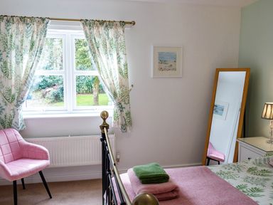 A bedroom with a bed with folded towels a pink chair a window with floral curtains and a full length mirror at The Old Vicarage Retreat in Luxulyan