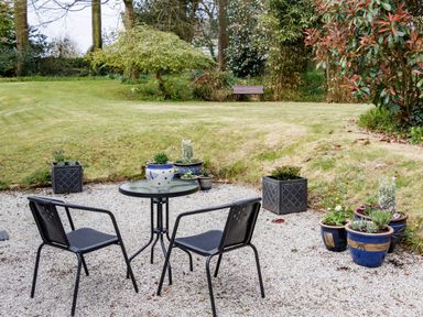 A garden patio with two chairs a round glass table and several potted plants on gravel with lawn and trees in the background at The Old Vicarage Retreat in Luxulyan