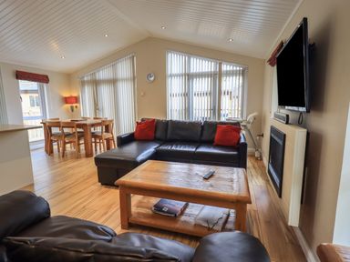 A living room with a black leather sofa and red cushions a wooden coffee table a dining table set and a wall-mounted television at Oakwood Lodge Langdale 3 White Cross Bay Windermere