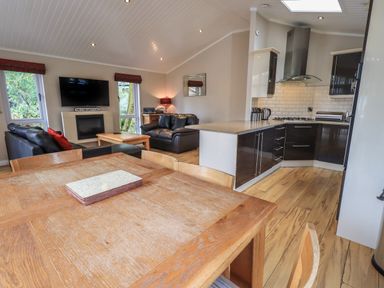 An open living room with wooden dining table, black leather sofas, a coffee table, a wall-mounted TV, and a modern kitchen at Oakwood Lodge Langdale 3 White Cross Bay Windermere