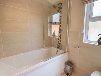 A bathroom with a bathtub shower combo a glass shower screen a window and a small trash bin at Oakwood Lodge in Langdale 3 White Cross Bay Windermere