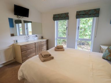 A bedroom with a bed, two towels on the bed, a dresser with a large mirror, windows with blue patterned curtains in Oakwood Lodge Langdale 3 White Cross Bay Windermere