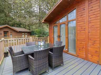 An outdoor wooden deck with a glass table and six wicker chairs in front of a cabin at Oakwood Lodge Langdale 3 White Cross Bay Windermere