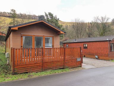 A wooden lodge with a fenced porch and French doors next to a similar lodge in a rural area at Lakeland View Lodge Beckside 2 Limefitt Park near Windermere