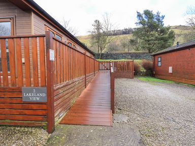 A wooden deck ramp leading to a cabin with a gravel driveway and trees in the background at Lakeland View Lodge in Beckside 2 Limefitt Park near Windermere