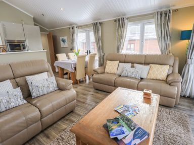 A living room with beige sofas, patterned cushions, a wooden coffee table with books and brochures, dining table with chairs near windows at Lakeland View Lodge Beckside 2 Limefitt Park near Windermere