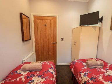 A bedroom with two single beds covered in red floral bedding towels on beds a wooden door a wall mirror a white wardrobe with a tv mounted above it at Lakeland View Lodge Beckside 2 Limefitt Park near Windermere