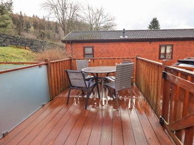 An outdoor wooden deck with a round glass table and four chairs and a wooden building in the background at Lakeland View Lodge Beckside 2 Limefitt Park near Windermere