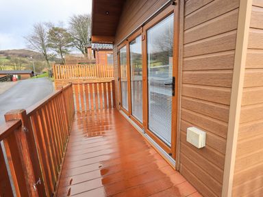 A wooden balcony with railing and glass doors of a cabin overlooking a road and trees at Lakeland View Lodge Beckside 2 Limefitt Park near Windermere
