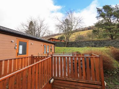 An outdoor wooden deck with chairs and table attached to a brown building with hills and trees in the background at Lakeland View Lodge Beckside 2 Limefitt Park near Windermere