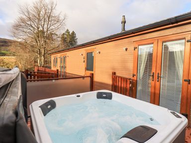 A hot tub on a wooden deck outside a lodge with trees and hills in the background at Lakeland View Lodge Beckside 2 Limefitt Park near Windermere