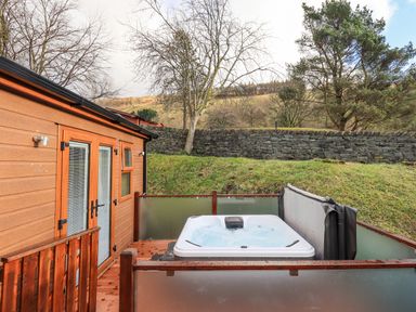 An outdoor hot tub on a wooden deck next to a wooden building with frosted glass panels and a stone wall in the background at Lakeland View Lodge in Beckside 2 Limefitt Park near Windermere