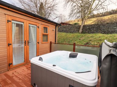 An outdoor hot tub on a wooden deck next to a building with wooden doors and windows at Lakeland View Lodge Beckside 2 Limefitt Park near Windermere