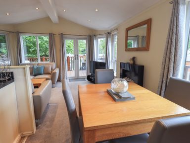 A dining table with chairs and a living area with sofas in a room with large windows and glass doors at Acorn Bank Lodge in L 55 Fallbarrow Park near Bowness-On-Windermere