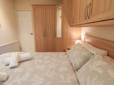 A bedroom with a bed, pillows, rolled towels, wooden wardrobes, and a door at Acorn Bank Lodge L 55 Fallbarrow Park near Bowness-On-Windermere