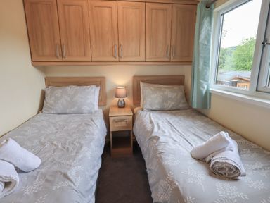 A bedroom with two single beds with patterned bedspreads and towels on them a nightstand with a lamp between the beds wooden cupboards above the headboards and a window with blue curtains at Acorn Bank Lodge L 55 Fallbarrow Park near Bowness-On-Windermere