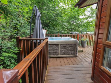 A wooden deck with a hot tub an umbrella and a potted plant surrounded by trees at Acorn Bank Lodge L 55 Fallbarrow Park near Bowness-On-Windermere