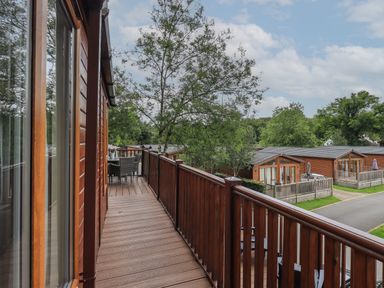 A wooden balcony with a table and chairs overlooking similar wooden lodges and trees at Acorn Bank Lodge L 55 Fallbarrow Park near Bowness-On-Windermere