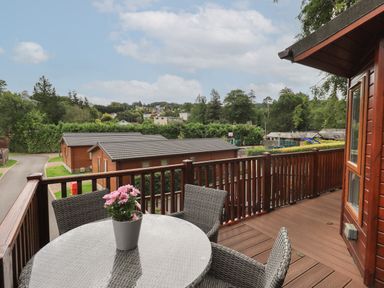 A balcony with a round table and chairs overlooking wooden lodges and trees at Acorn Bank Lodge in L 55 Fallbarrow Park near Bowness-On-Windermere