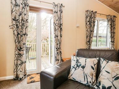 A living room with a leather sofa and floral patterned curtains by glass doors and a window at Damson Lodge Thirlmere 8 White Cross Bay Windermere