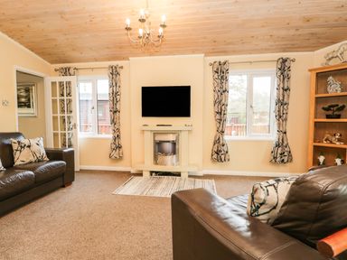 A living room with two brown leather sofas a wall-mounted television and fireplace two windows with patterned curtains and a wooden bookshelf at Damson Lodge Thirlmere 8 White Cross Bay Windermere
