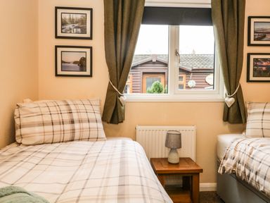 A bedroom with two beds with plaid bedding a wooden side table and lamp under a window with curtains at Damson Lodge Thirlmere 8 White Cross Bay Windermere