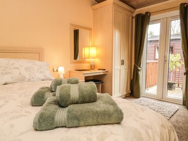 A bedroom with a bed with folded towels on it a desk with a lamp and mirror a wardrobe and glass doors with curtains at Damson Lodge Thirlmere 8 White Cross Bay Windermere