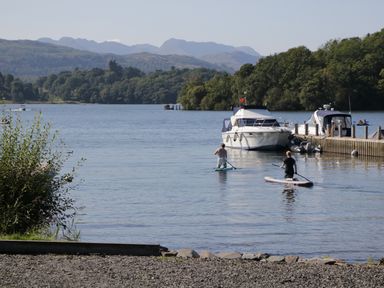 Two people paddleboarding near boats docked at a pier on a lake with trees and mountains in the background at Damson Lodge in Thirlmere 8 White Cross Bay Windermere