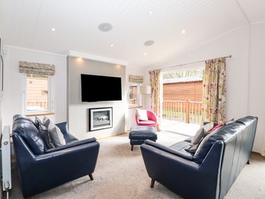 A living room with two blue sofas a pink chair a wall mounted television and floral curtains at Ridgway Lodge Calgarth 40 White Cross Bay near Windermere