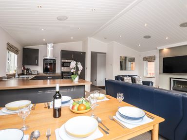 An open kitchen and dining area with a wooden table set with dishes and glasses a fruit bowl and wine bottle a living room with blue sofas a wall-mounted TV and a fireplace at Ridgway Lodge Calgarth 40 White Cross Bay near Windermere