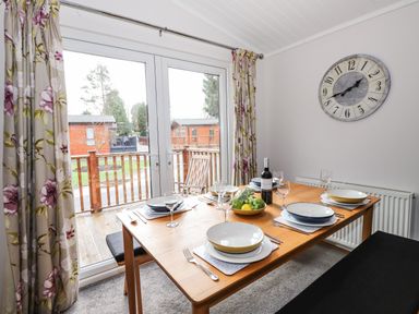A dining room with a wooden table set for four with plates wine glasses and a bowl of fruit near glass doors and floral curtains at Ridgway Lodge Calgarth 40 White Cross Bay near Windermere