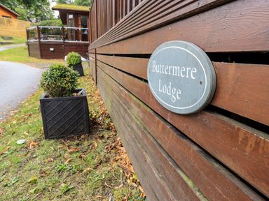 A wooden building with a sign reading Buttermere Lodge and potted bushes outside at Buttermere Lodge in Glade 5 Fallbarrow Park near Bowness on Windermere
