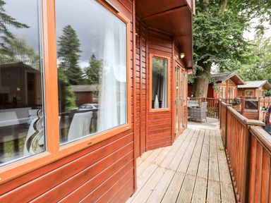 A wooden deck with railings outside cabins with large windows and a small table with chairs at Buttermere Lodge in Glade 5 Fallbarrow Park near Bowness on Windermere