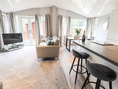 A living room with a sofa, television, dining table, and bar stools at Buttermere Lodge in Glade 5, Fallbarrow Park near Bowness on Windermere