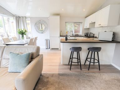 An open plan living area with a leather sofa dining table with chairs kitchen island with two stools and kitchen cabinets at Buttermere Lodge in Glade 5 Fallbarrow Park near Bowness on Windermere
