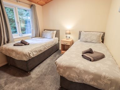 A bedroom with two single beds a nightstand with a lamp between them and a window with curtains at Buttermere Lodge Glade 5 Fallbarrow Park near Bowness on Windermere